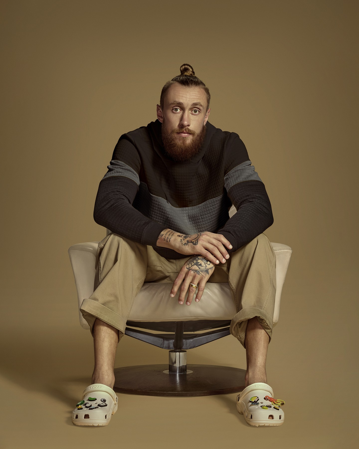 Bearded man with hair bun sits in white chair against tan background. He wears a striped sweater, khaki pants, and clogs. Tattoos visible.